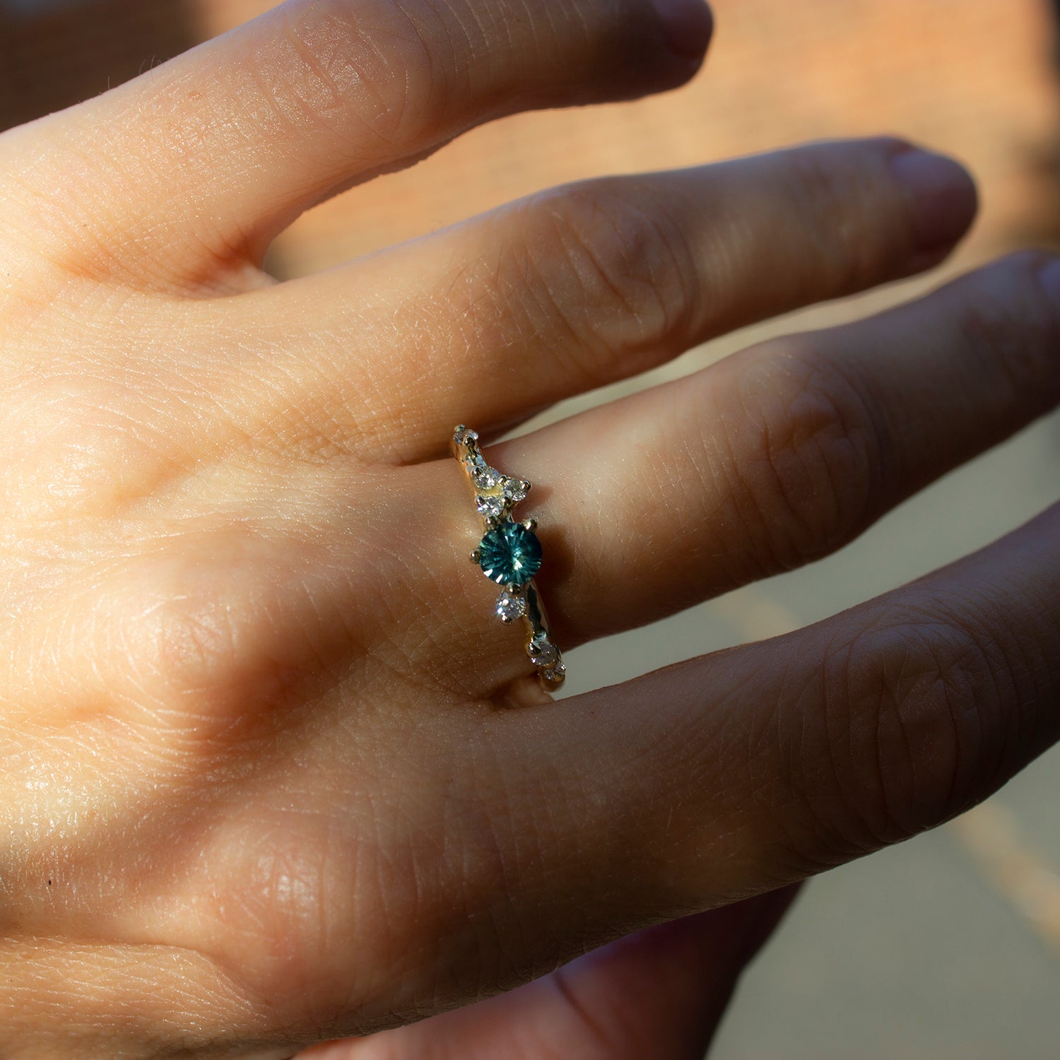 Asymmetrical, delicate ring featuring a beautiful teal sapphire and white diamonds scattered along a gold band.